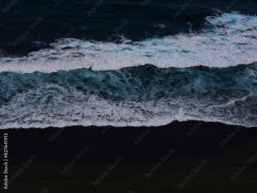 Fototapeta premium Aerial photo of a sea water surface. Blue water with foam on waves. Beach with black volcanic sand - view from above. Stylized background abstract texture photo. Tenerife, Puerto de la Cruz
