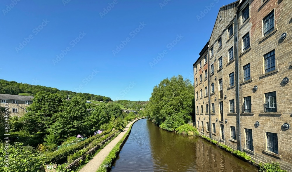 The Rochdale canal runs beside a former Victorian mill, with windows, reflecting a clear blue sky. old trees, and greenery, create a picturesque setting in, Todmorden, UK The Rochdale canal runs beside a former Victorian mill, with windows, reflecting a clear blue sky. old trees, and greenery, create a picturesque setting in, Todmorden, UK
