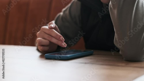 A woman uses her smartphone, gently scrolling with her finger. The blue phone rests on a warm-toned wooden table in a cafe, creating a smooth and detailed video at 50fps. photo