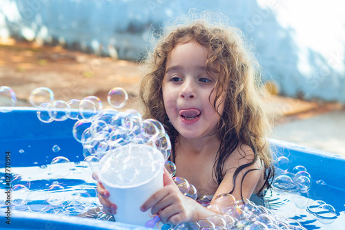 Uma garotinha dentro de uma piscina infantil azul, brincando com bolhas de sabão no quintal de casa. photo