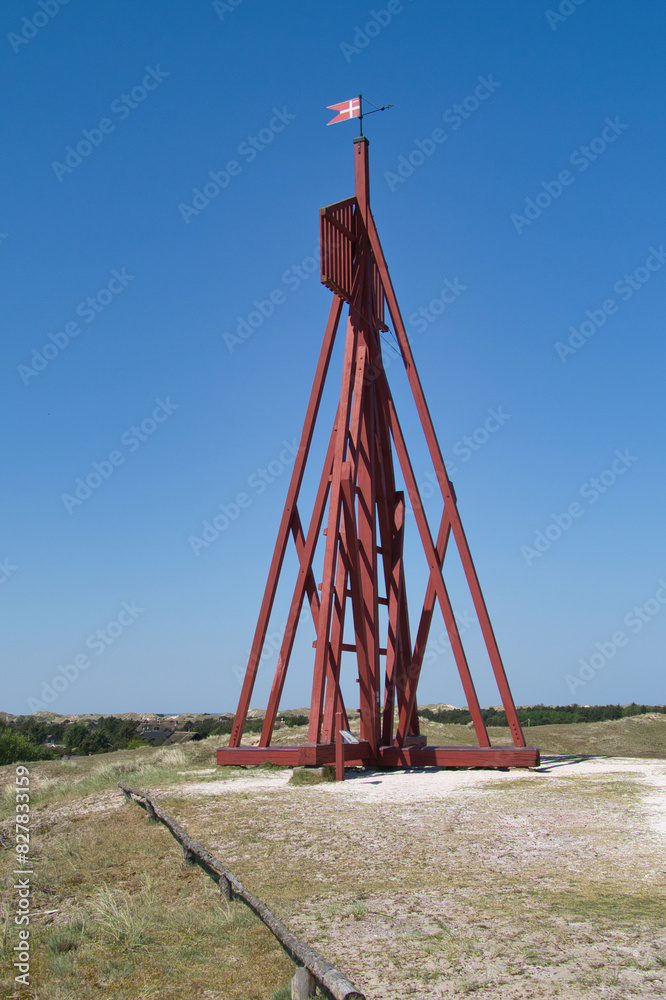 A tall, red wooden navigation mark stands against a clear blue sky. The structure, featuring a Danish flag on top, is set in a coastal landscape with sandy ground and sparse vegetation A tall, red wooden navigation mark stands against a clear blue sky. The structure, featuring a Danish flag on top, is set in a coastal landscape with sandy ground and sparse vegetation