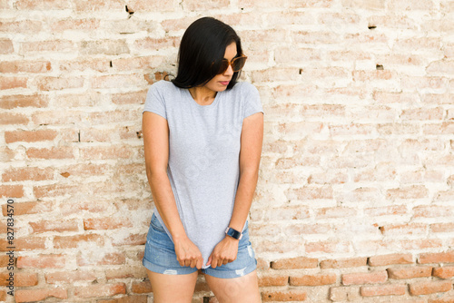 Portrait of a young latina woman in a plain grey t-shirt, posing against a weathered brick wall for branding purposes