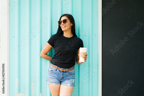 Confident latin woman in black t-shirt holding coffee cup stands against teal background with sunglasses on a sunny day, smiling