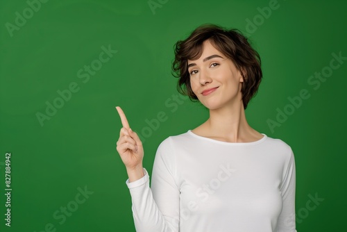 short haired young happy women wearing white long sleeved top pointing upwards with their right index finger against solid green background copy space