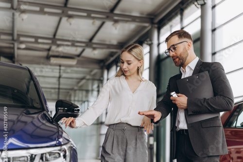 Car Sales Manager Showing Auto To Caucasian Lady Buyer Standing In Luxury Automobile Dealership Store. Buying Vehicle Concept
