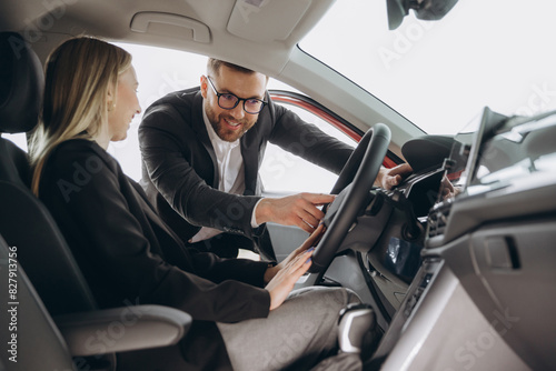 Friendly male manager showing young female customer cars. Beautiful smiling woman sitting inside vehicle in car. Bearded sales consultant working in car dealership.