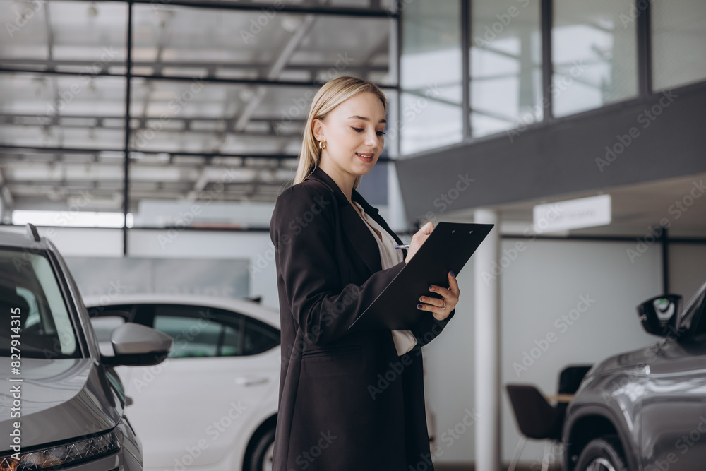 Handsome car dealership female worker in suit is holding a folder and smiling while standing near the car Handsome car dealership female worker in suit is holding a folder and smiling while standing near the car
