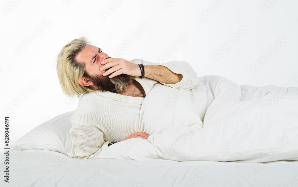 Handsome man in bathrobe yawning waking up in cozy bed at home in morning. Sleepy man in bed at bedroom yawning covering mouth with palm. Bearded man yawning lying on soft white pillow. Yawn and nap. Handsome man in bathrobe yawning waking up in cozy bed at home in morning. Sleepy man in bed at bedroom yawning covering mouth with palm. Bearded man yawning lying on soft white pillow. Yawn and nap.