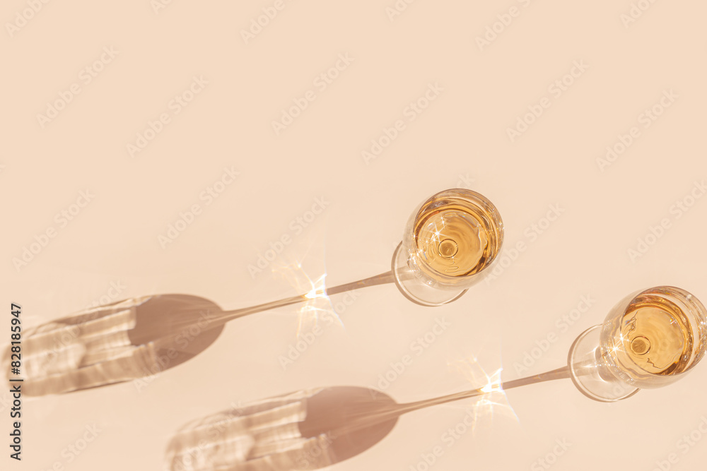Sunlight still life with two glasses of white wine, beige background with shadow and glare, caustic light effect at sun. Minimal aesthetic trend photo with summer alcohol drink, dark light, above