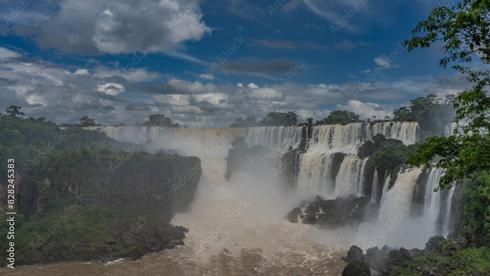 A beautiful cascade of waterfalls. White foaming streams collapse from the rocks into the riverbed. Spray, fog rising. Clouds in the blue sky. Lush green vegetation. Iguazu Falls. Argentina. A beautiful cascade of waterfalls. White foaming streams collapse from the rocks into the riverbed. Spray, fog rising. Clouds in the blue sky. Lush green vegetation. Iguazu Falls. Argentina.