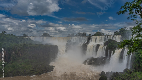 A beautiful cascade of waterfalls. White foaming streams collapse from the rocks into the riverbed. Spray  fog rising. Clouds in the blue sky. Lush green vegetation. Iguazu Falls. Argentina.