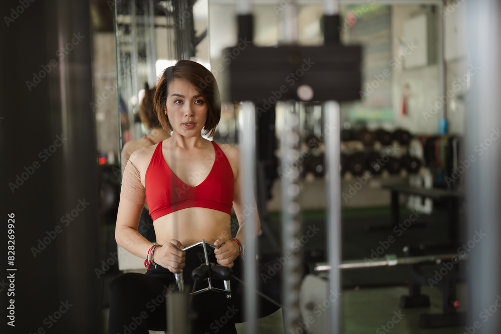 Fototapeta premium Sport determined woman in a red sports bra and black leggings uses a seated row machine in a modern gym. Her focused expression and fit physique emphasize her commitment to strength training.