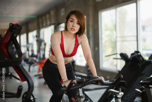 Sport woman in a red sports bra and black leggings rides a stationary bike in a modern gym. Her determined expression and fit physique highlight her dedication to fitness.