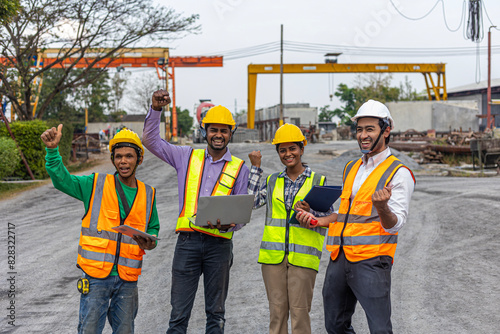 Team of civil engineers from different cultures celebrate their achievement by putting hand up. Workers in safety vests using laptop or tablet to monitor their production in a construction factory..