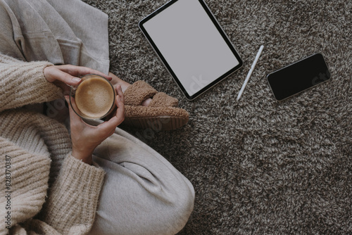 Cozy morning routine. Top view of woman wearing pastel creamy beige clothes sitting on fluffy grey carpet with cup of coffee. Tablet and mobile phone mock up with copy space © Floral Deco