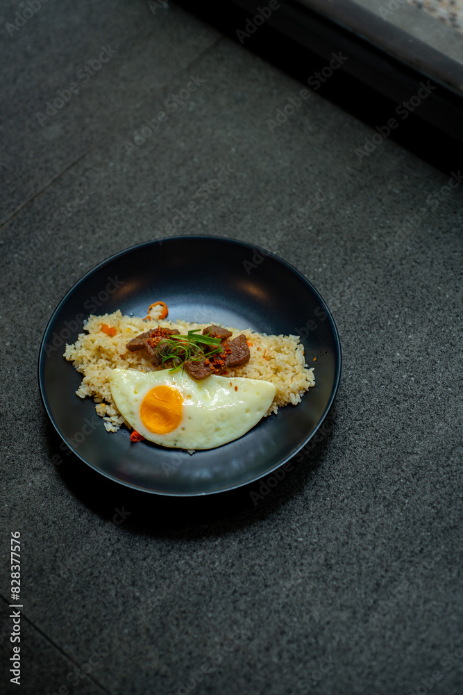 beef saikoro rice bowl and sunny side up egg on black plate, over stone tile