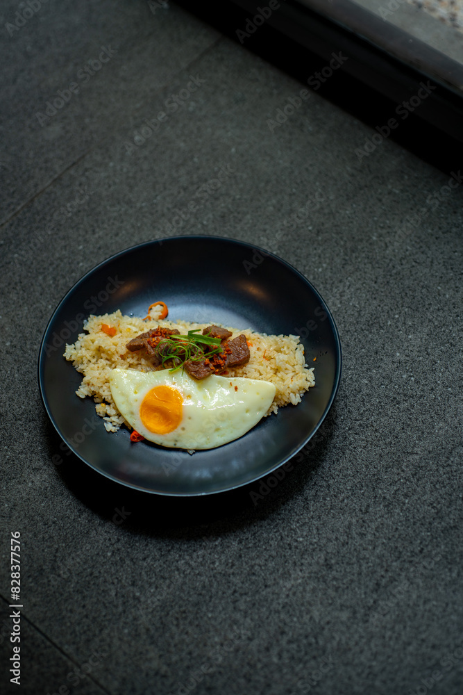 beef saikoro rice bowl and sunny side up egg on black plate, over stone tile beef saikoro rice bowl and sunny side up egg on black plate, over stone tile