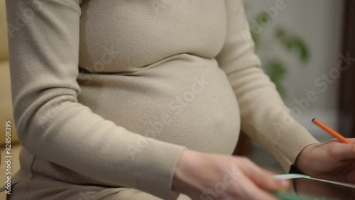 Close-up. An unrecognizable pregnant woman works remotely from home. A woman holds an open tablet in her hands and compares data while sitting in a leather armchair in the living room at home