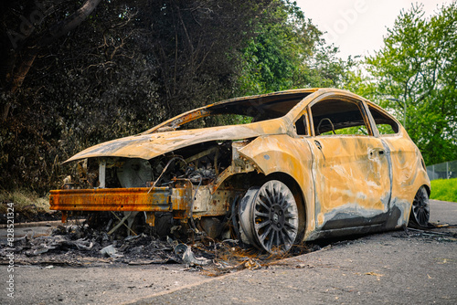  burnt out car abandoned on concrete footpath rusty and orange colour with hedgerows in the background 