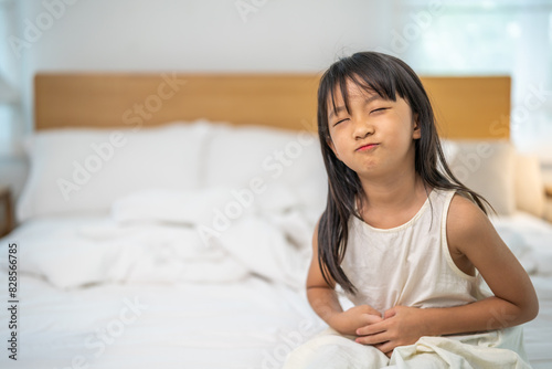 Portrait of sick unhappy little child asian girl touching belly stomach with suffering from stomach ache pain, gastritis, gastroenteritis, food poisoning, diarrhea, intestinal inflammation at home