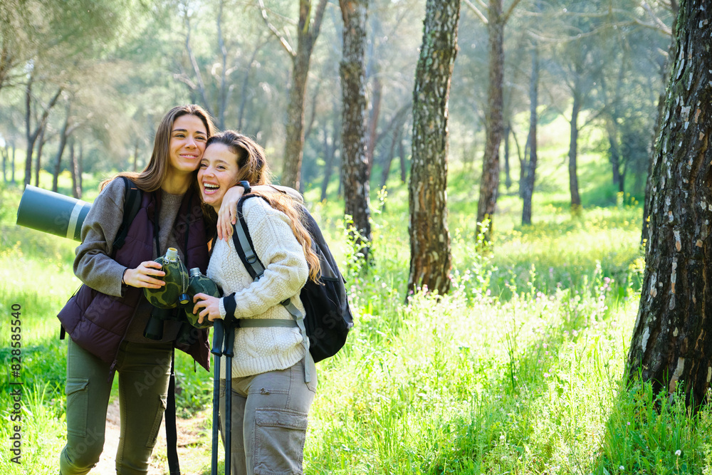 Two women are smiling and hugging each other while holding water canteen. They are doing trekking or hiking in a forest