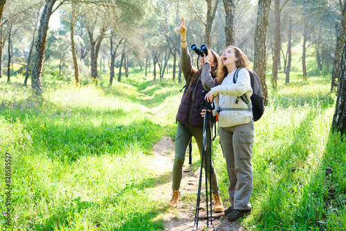 Two women are looking through binoculars in a forest. One of them is pointing at something in the distance