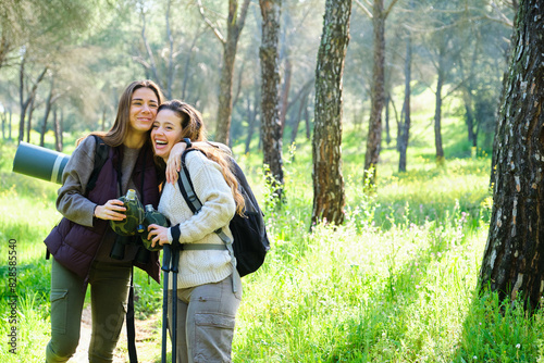 Obraz na płótnie Two women are smiling and hugging each other while holding water canteen. They are doing trekking or hiking in a forest w salonie