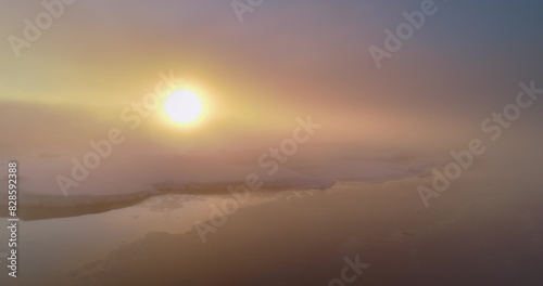 Aerial fly over sunrise fog sky in Antarctica. Morning sun shine through clouds on snow covered hill mountains ocean. Epic panorama overview, bright orange sun rising up. Nature travel background