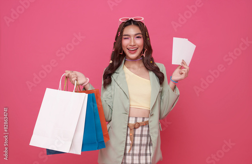 Happy pretty Asian woman carrying colorful shopping bags with gift voucher flyer mock up isolated on pink studio background.