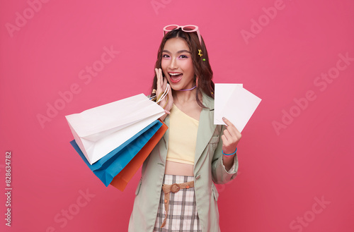 Happy pretty Asian woman carrying colorful shopping bags with gift voucher flyer mock up isolated on pink studio background.