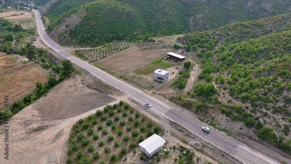 Aerial view of cars driving on a highway through green, hilly farmland with small buildings and cultivated fields