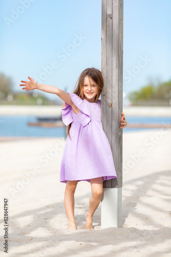 Happy little blonde girl in lilac summer dress playing and has fun on a white sandy beach near the ocean in summer. Children's holidays. Earth Day. Children's Day. Vacation concept. Space for text