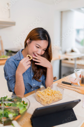 Young asian woman eating snack and watching movie entertainment on tablet after preparing fresh vegetables salad and cooking healthy breakfast food in modern kitchen with healthy lifestyle at home
