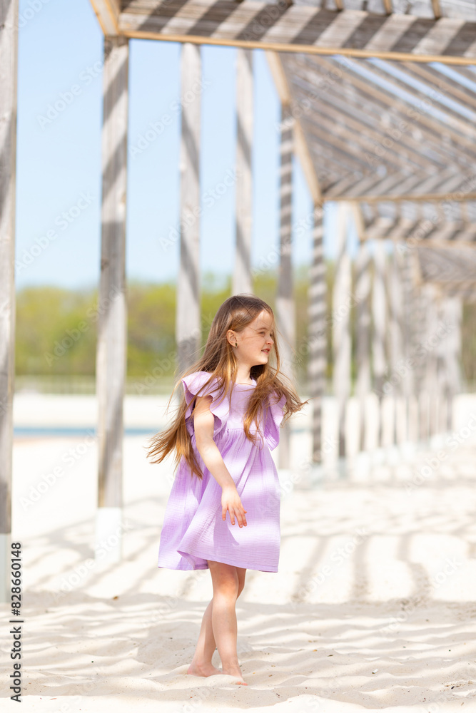 Happy little blonde girl in lilac summer dress playing and has fun on a white sandy beach near the ocean in summer. Children's holidays. Earth Day. Children's Day. Vacation concept. Space for text