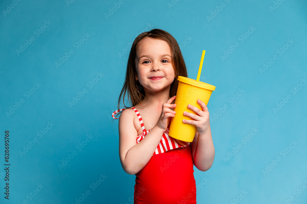 Little smiling beautiful girl in red swimsuit drinking lemonade through a tube on blue background, Summer, beach concept. Copy space. Little smiling beautiful girl in red swimsuit drinking lemonade through a tube on blue background, Summer, beach concept. Copy space.