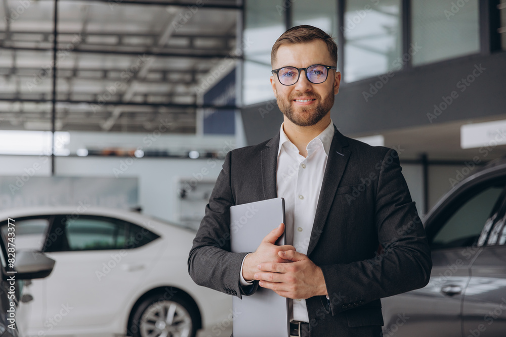Modern bearded man in glasses and suit vehicle sales consultant using laptop inside car dealership Modern bearded man in glasses and suit vehicle sales consultant using laptop inside car dealership