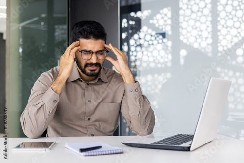 In the office, a midadult caucasian man is visibly stressed while working at his desk with a laptop, surrounded by documents, pens, and a notepad. He appears focused on his tasks photo