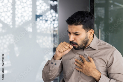 The image depicts a man in discomfort while holding his chest and coughing in a workplace setting. Keywords related to the scene include illness, stress, symptoms, and respiratory problems