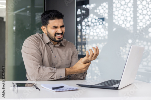 A businessman looks uncomfortable using his laptop in a techfocused office, showing the strain of repetitive tasks and emphasizing the pressure and stress in such environments photo