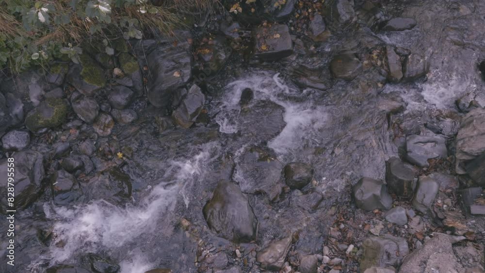 Small stream flowing over rocks and pebbles surrounded by greenery. Water creates white foam as it moves through rocky terrain. Small stream flowing over rocks and pebbles surrounded by greenery. Water creates white foam as it moves through rocky terrain.