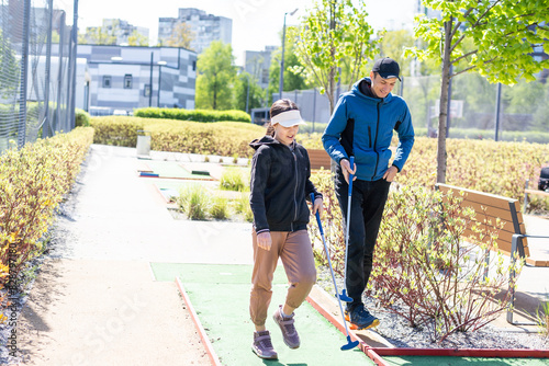 The girl is preparing to hit the ball with a golf club and looks at the man who is squatting next to her with the golf club in his hands.