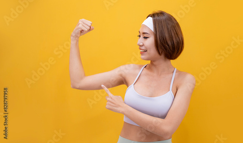 Portrait of healthy and slim Asian athlete woman in sportswear smiling and pointing on bicep isolated on yellow background for exercise and workout concept