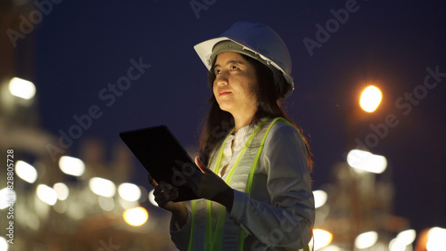 A female engineer wearing a helmet and reflective vest multitasks with a tablet and phone at a brightly lit industrial site at night.