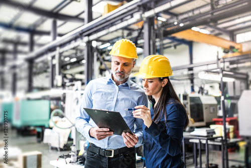 Two project managers standing in modern industrial factory. Manufacturing facility with robotics, robotic arms and automation. Storing products and materials in warehouse.