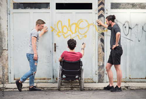 Young man in wheelchair spending free time outdoor with friends, drawin on abadoned building with spray paint. Male friendship. photo
