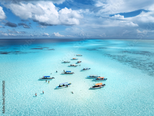 Aerial view of colorful boats in clear azure water in summer. Mnemba island, Zanzibar. Top drone view of sandbank in low tide, blue sea, white sand, swimming people, yachts, sky with clouds. Ocean photo