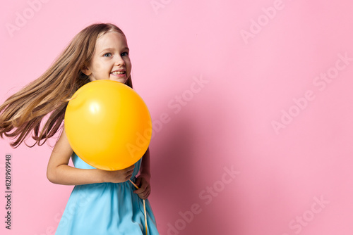 Little girl in a blue dress joyfully holding a yellow balloon against a vibrant pink background © SHOTPRIME STUDIO