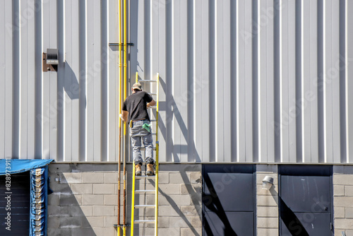 Man standing on a ladder painting natural gas lines yellow on the side of a building with grey vertical siding, daytime, sunshine, nobody