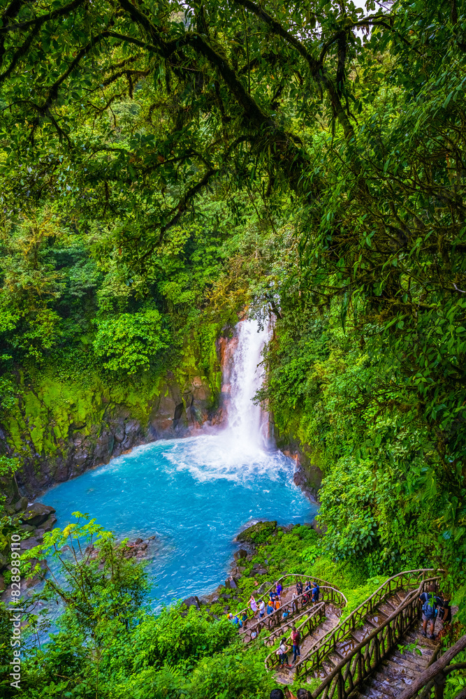 Fototapeta premium A vibrant waterfall cascades into a turquoise pool surrounded by lush green rainforest. A group of tourists descends a rustic staircase to view the natural beauty up close. High quality photo. Rio
