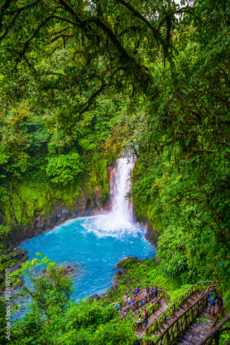 A vibrant waterfall cascades into a turquoise pool surrounded by lush green rainforest. A group of tourists descends a rustic staircase to view the natural beauty up close. High quality photo. Rio